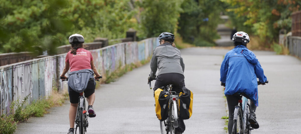 3 cyclists, wearing helmets, cycling over bridge in Avenham Park