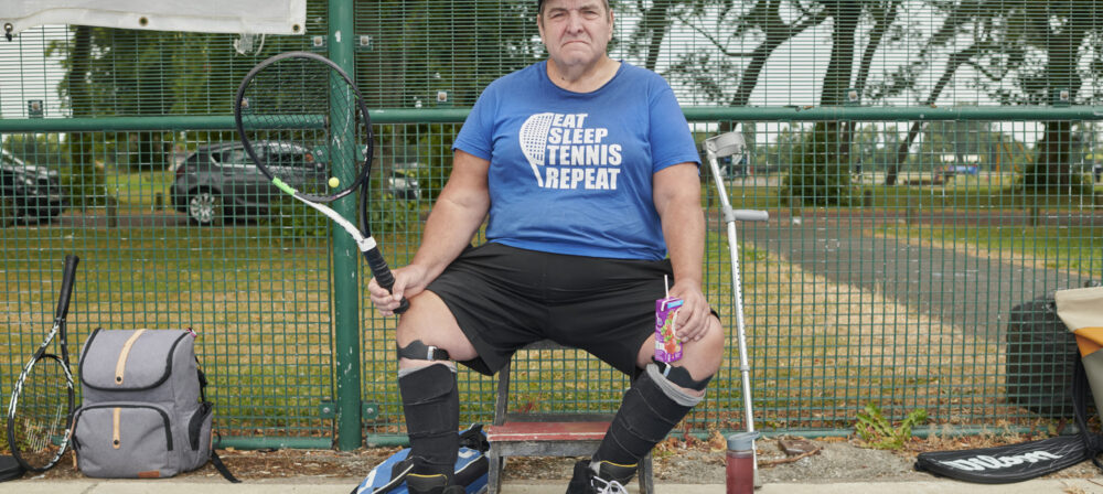 Male sitting next to net on tennis court, holding racket
