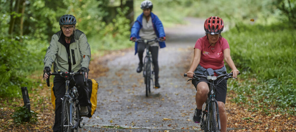 Three female cyclists, wearing helmets, cycling on path through trees