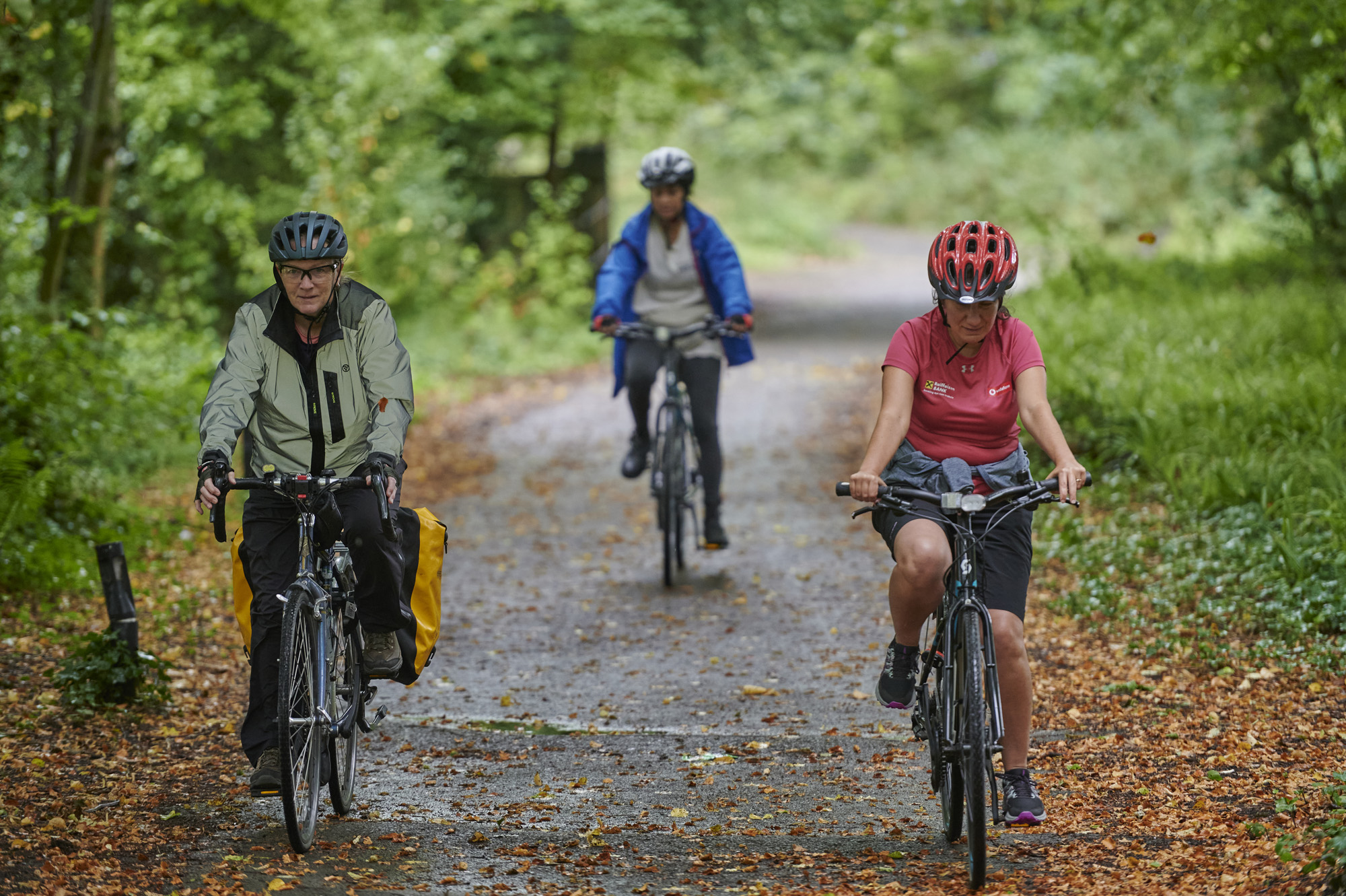 Three female cyclists, wearing helmets, cycling on path through trees