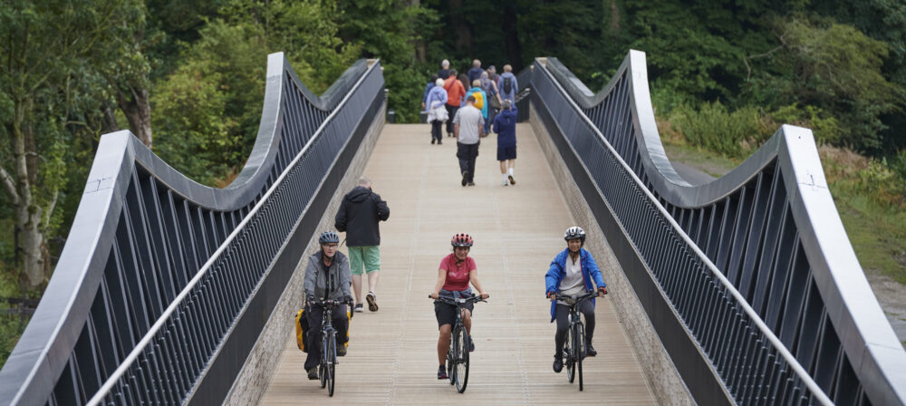Three cyclists, wearing helmets, cycling over Tram Bridge, at a distance