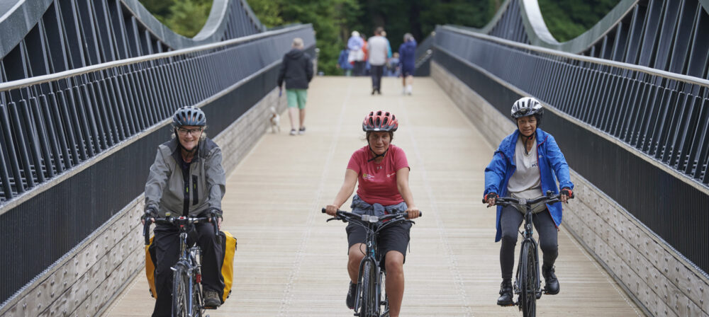Three female cyclists, wearing helmets, cycling across Tram Bridge towards the camera