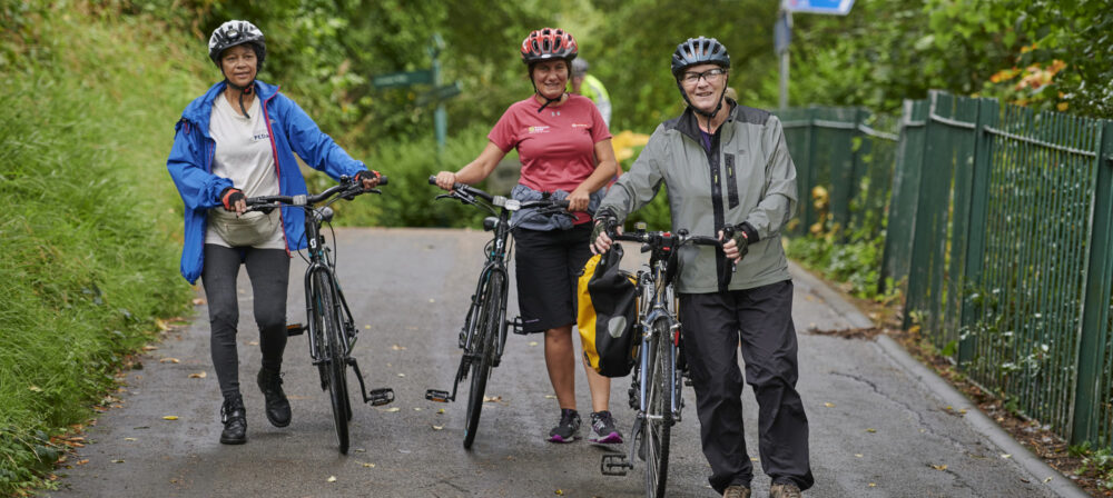 Three female cyclists, wearing helmets, standing with their bikes