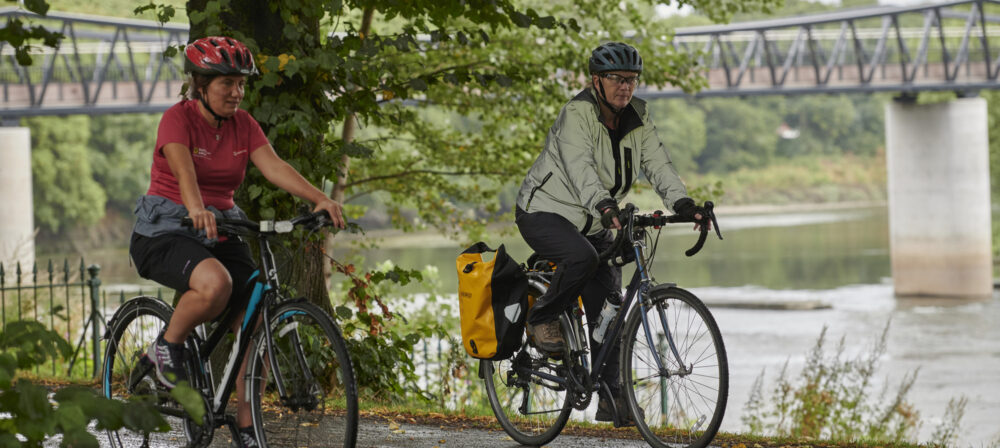 Two female cyclists, wearing helmets, cycling down path