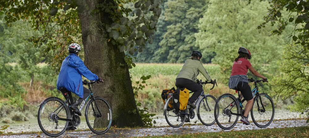Three females, wearing cycling helmets, cycling through countryside