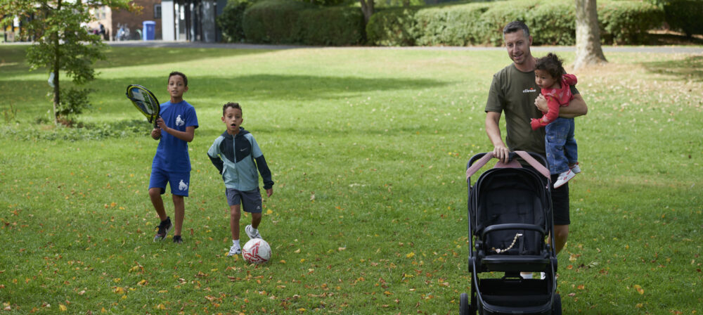 Father holding baby daughter whilst pushing the pram, two brothers walking beside the pram