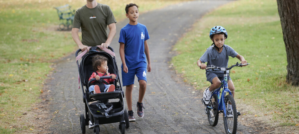Male pushing pram with young boy walking beside him and younger boy on bike wearing helmet