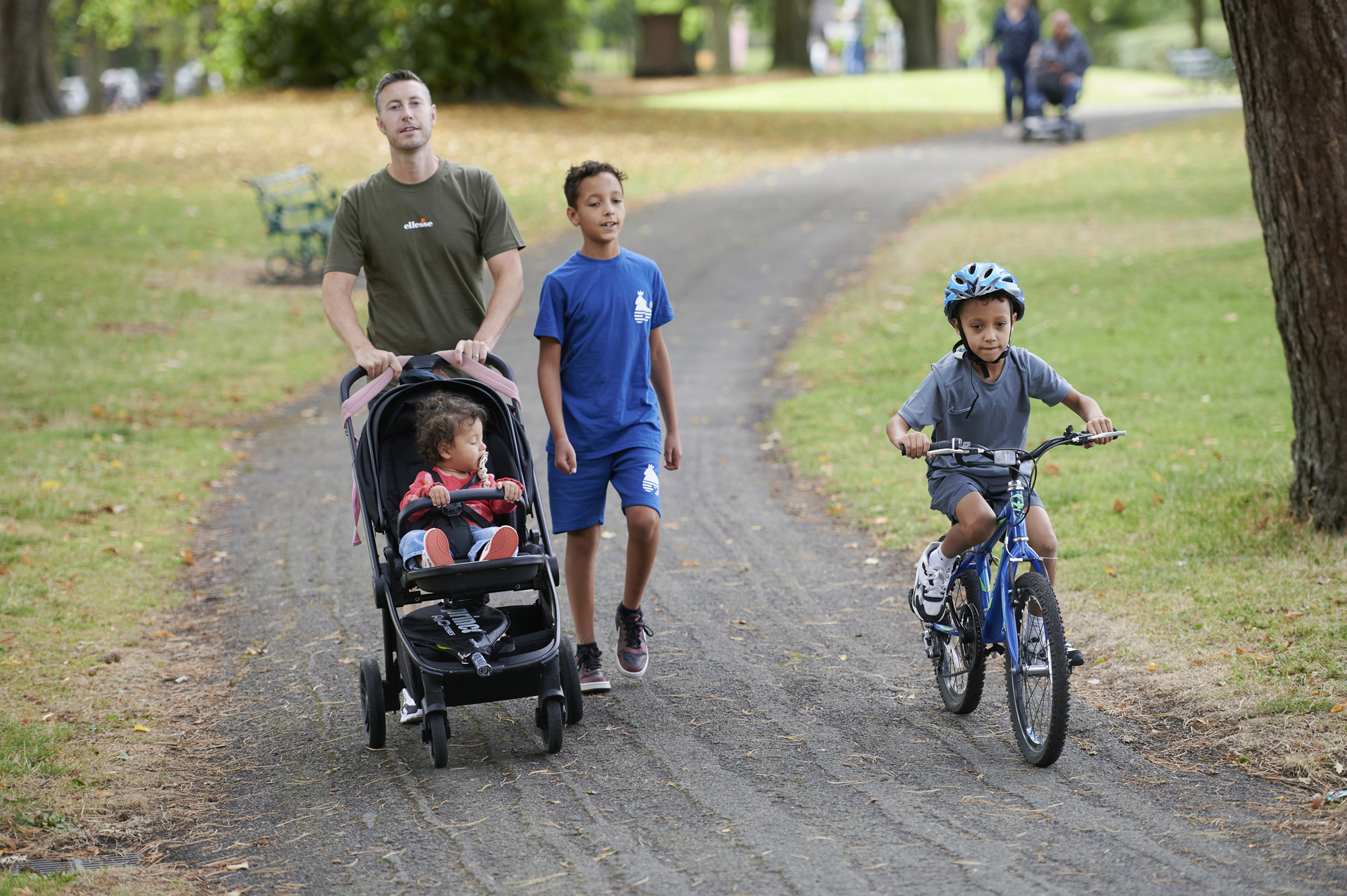 Male pushing pram with young boy walking beside him and younger boy on bike wearing helmet