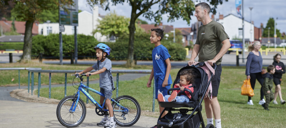 Male pushing pram with young girl, one boy scottering and one boy walking